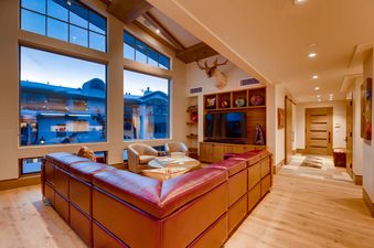 A luxurious living room in Gondola One Chalet featuring a large red leather sectional sofa, a flat-screen TV with custom shelving, and expansive floor-to-ceiling windows offering a snowy exterior view.