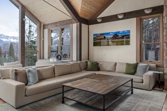 The living area features a large sectional sofa, a rustic coffee table, and expansive windows offering a scenic view of mountains and trees.