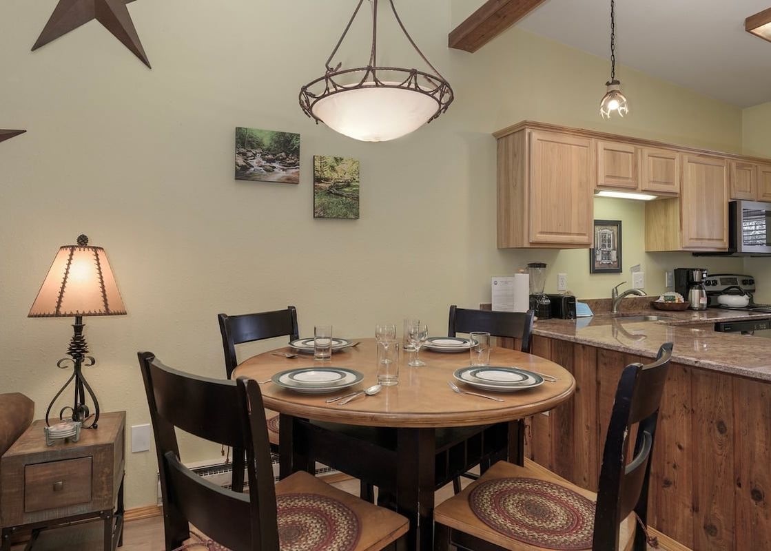 A cozy dining area features a round wooden table set for four, with a large overhead chandelier and rustic decor elements.