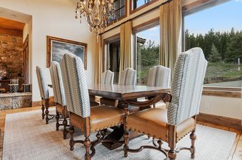 The dining area at The Last Best Chalet features a large wooden table, elegant chairs with leather and patterned fabric, a grand chandelier, vaulted wooden ceilings, and expansive windows showcasing outdoor views.