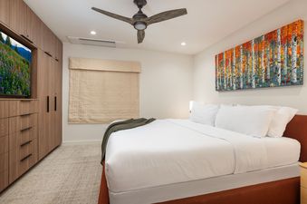 A modern bedroom with a king-size bed, wood cabinetry with a television, and a colorful aspen tree painting.