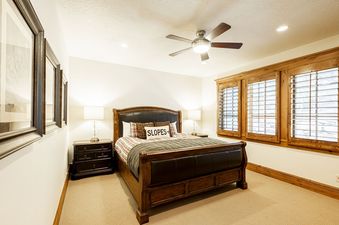 The bedroom features a dark wooden sleigh bed with a brown leather headboard and footboard, flanked by matching nightstands with lamps, and two large windows with wooden shutters.