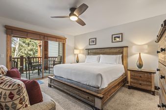 A spacious and inviting primary bedroom featuring a king-size bed, rustic wooden furniture, soft lighting, and access to a private balcony with outdoor seating and a view of green trees.