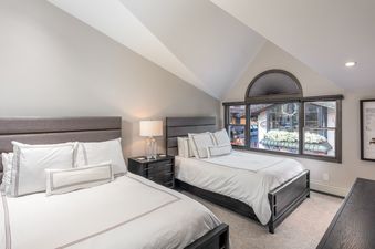 A cozy bedroom with two beds, each adorned with crisp white bedding and gray accent stripes. The room features a sloped ceiling and a large window offering a view of an outdoor street with shops and white flowers in window boxes.