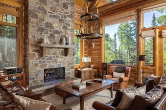 The living room at Bear Lodge features a large stone fireplace, leather seating, a wooden coffee table, and windows showcasing a forest view.