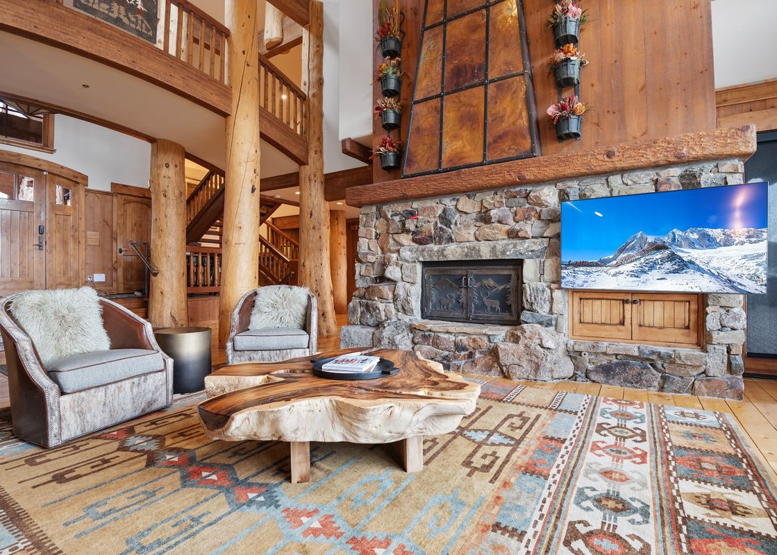 A spacious and luxurious living room at the Timber Lodge at the Oaks, featuring a large stone fireplace, a flat-screen TV, two armchairs with faux fur pillows, and a unique wooden coffee table.