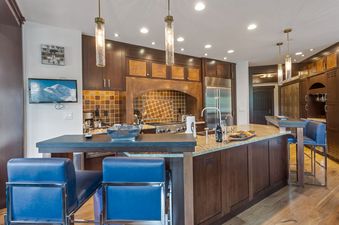 A modern kitchen with dark wood cabinets, a granite island, stainless steel appliances, and blue bar stools, illuminated by pendant and recessed lighting.