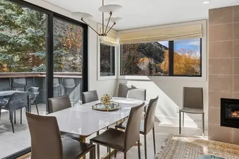 A modern dining room with a white rectangular table and dark grey chairs, featuring large windows with views of autumn trees and mountains.