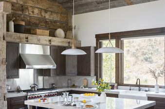A rustic kitchen at Northern Exposure featuring a large white marble island with dark blue bar stools, dark wood cabinetry, stainless steel appliances, and exposed wooden beam ceilings.
