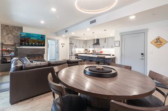 A modern and inviting open-concept living space in The Aerie, featuring a large television above a fireplace, a comfortable leather sectional, and a circular dining table, all seamlessly connected to a contemporary kitchen.
