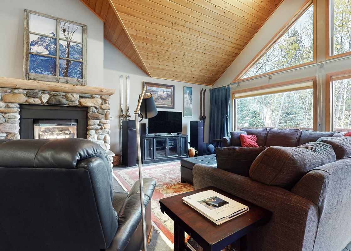 A cozy living room with a large stone fireplace, comfortable sofas, and big windows revealing a view of trees outside.