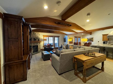 A spacious and inviting living area inside The Chateau Deluxe Four Bedroom Suite, featuring a large grey sectional sofa, a stone fireplace, and a wooden entertainment unit.