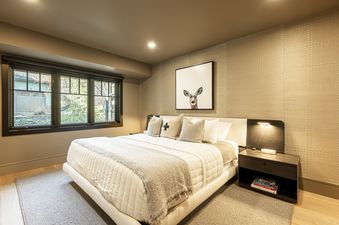 The primary bedroom at Weedon Ranch features a large bed with neutral bedding, a decorative accent wall, a bedside table with a lamp, and a window offering a view of the outdoors.