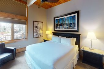 The bedroom of Manor Vail 382 features a king-size bed with a white duvet and a large window with a view of the trees outside.