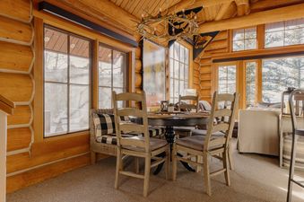 A rustic dining area with a round black table, wooden chairs, and a wicker chair with buffalo check cushions, set within a log cabin interior with large windows looking out onto a winter landscape.