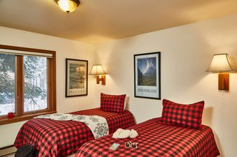 A cozy bedroom in the Teewinot B3 unit with two twin beds featuring red and black plaid bedding, wall sconces, framed travel posters, and a window looking out onto snowy trees.