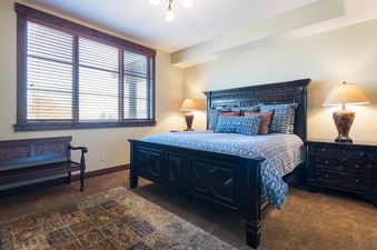 A spacious bedroom features a large dark wooden bed with ornate carvings, blue and rust-colored bedding, and two matching nightstands with lamps, all set on a brown carpet.