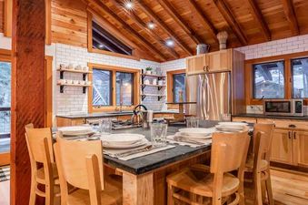 A kitchen with a large dining table and chairs, a stainless steel refrigerator, and a view of the trees outside.
