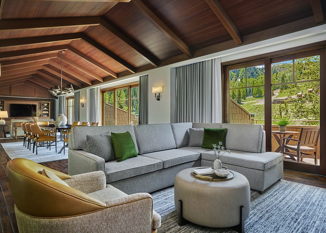 A spacious Montage Flagstaff Suite living area with a large grey sectional sofa, an ottoman, and an armchair, facing large windows with an exterior view of a lush green hillside.