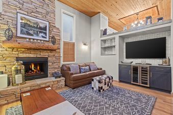 The inviting lounge area at Motherlode 3 features a stone fireplace, a brown leather sofa, and a built-in wet bar with a TV, all under a warm wooden ceiling.