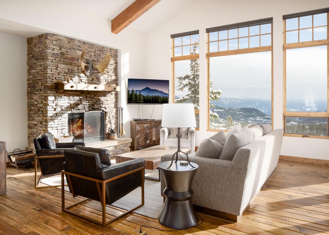 A luxurious living room with a stone fireplace, smart TV, comfortable seating, and large windows revealing a stunning snowy mountain landscape.