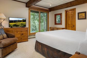 A bedroom with a white bed with a brown leather headboard, and a large window with a view of trees.