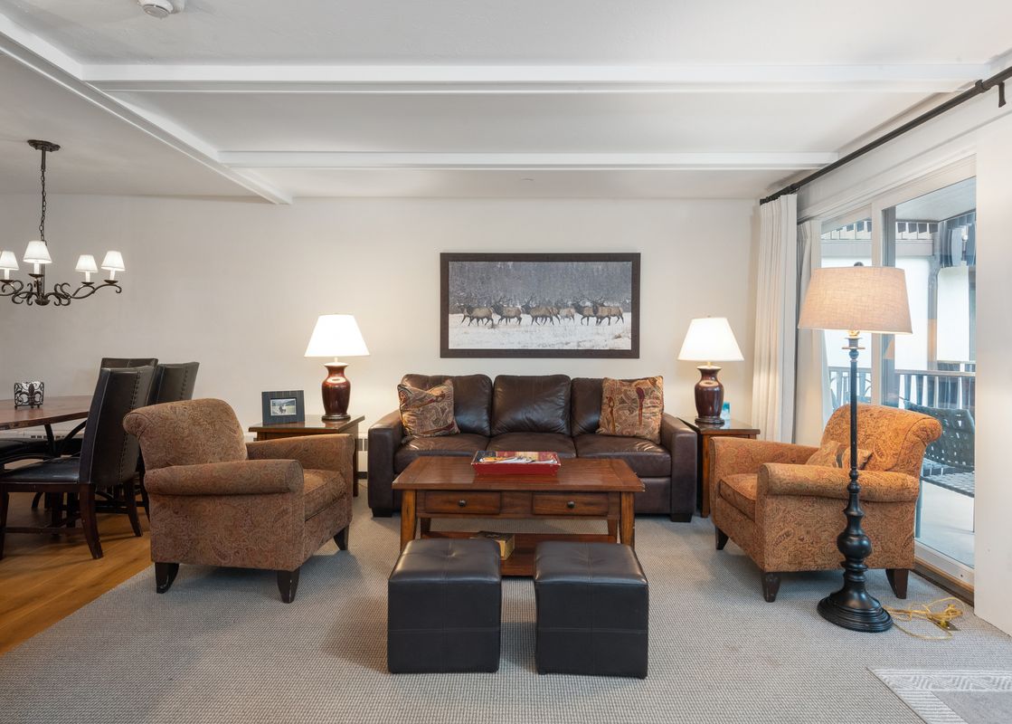 The living area features a large brown leather sofa, two patterned armchairs, a wooden coffee table, and two black ottomans, with a dining area visible in the background and natural light from a large window.