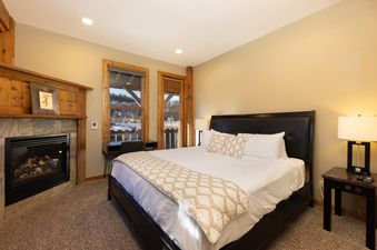 A cozy bedroom in The River House Lodge features a large bed with white and patterned bedding, a fireplace with a stone surround, and windows offering a view of a snowy landscape.