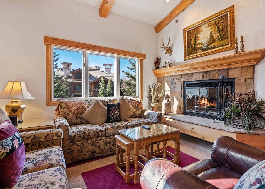 A cozy living room at The Charter E2205 featuring a stone fireplace, comfortable floral sofas, a wooden coffee table, and a large window overlooking other village buildings and pine trees.