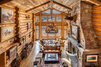 A high-angle view of the rustic, spacious great room in Elk Mountain Lodge, featuring log walls, a vaulted ceiling, large windows with mountain views, and a grand antler-themed chandelier.