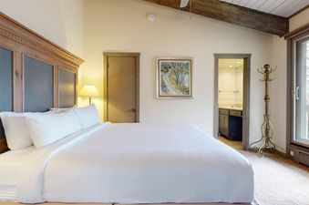 A cozy bedroom at Top of the Village Leaf 503 features a large bed with white linens, a wooden headboard, a bedside lamp, and a painting of a snowy road, with an adjacent bathroom and a window offering an exterior view.