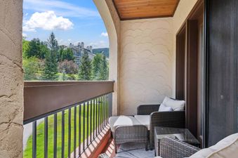 A private balcony with wicker lounge chairs and a glass-top table, offering views of green trees and distant buildings under a blue sky.