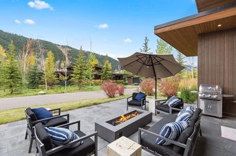A modern outdoor patio at the Fairway Cabin Two Bedroom features a fire pit, comfortable seating with blue and striped pillows, and a BBQ grill, all set against a backdrop of lush trees and mountains under a blue sky.