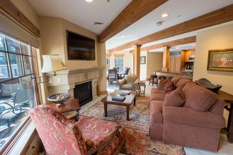 The Chateaux Luxury One Bedroom boasts a spacious and inviting lounge area with a fireplace, comfortable seating, and a large window offering an exterior view of lush greenery.