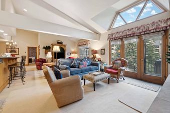 A cozy and inviting living room at The Charter E430 featuring a blue sofa, armchairs, and large windows with views of evergreen trees, all under a vaulted ceiling with skylights.