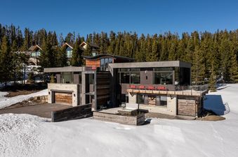 A modern multi-level house, The View, stands amidst a snow-covered landscape and towering pine trees under a clear blue sky, featuring a hot tub on an outdoor deck.