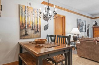 A rustic dining area with a dark wood table and chairs, featuring a large painting of birch trees on the wall and a classic chandelier overhead.