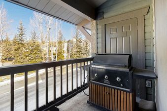 A black barbecue grill sits on the balcony of Phoenix 216, surrounded by a metal railing and offering a view of snow-dusted evergreen and bare deciduous trees, with a building visible in the background under a blue sky.