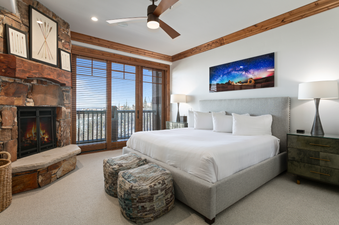 A master bedroom in Grand Lodge Three Bedroom Deluxe 408 features a large king-size bed, two plush ottomans, and a view to the surrounding mountains through the window.