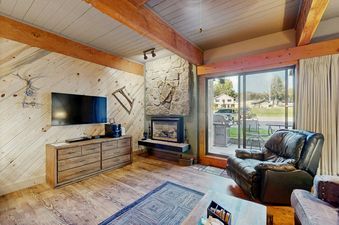 A cozy living room with wood-paneled walls, a fireplace, a large TV, and a leather armchair, with a sliding glass door leading to an outdoor patio.