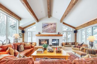 A cozy, spacious living room with rustic wooden beams on the ceiling, a large fireplace, comfortable sofas, and expansive windows offering a view of a snowy forest.