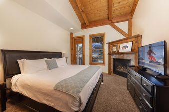 The bedroom at The River House Lodge features a comfortable bed with a dark headboard, a large fireplace to the right, and a flat-screen TV on a dresser, with large windows providing a view of the snow-covered outdoors.