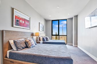 A modern bedroom with two queen beds, each with blue bedding and a mix of pillows, a nightstand with a lamp, a wall-mounted TV, and a large window offering an exterior view of trees and sky.