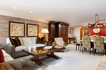 A spacious living and dining area features a light-colored sectional, a glass-top coffee table on a cowhide rug, and an elegant dining table with paisley upholstered chairs under a chandelier.