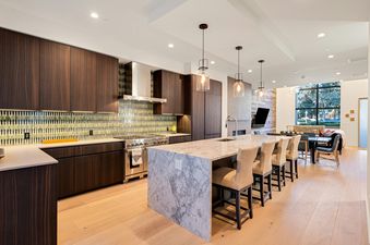 A modern kitchen and living area with dark wood cabinets, a mosaic tile backsplash, a marble island with bar stools, and a large window looking out to trees.