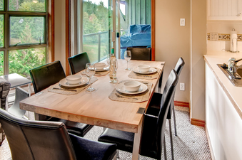 The dining area in Aspens on Blackcomb Unit 552 features a large wooden table set for six with dinnerware and wine, complemented by black chairs and a view of the forest outside.