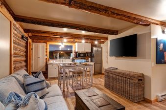 A living room with a couch, a coffee table, a large screen TV, and a wicker storage bench. The room is decorated with rustic wood beams and a log cabin wall.