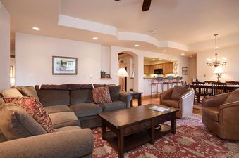 A spacious and inviting living room featuring a large brown sectional sofa with patterned throw pillows, a dark wood coffee table, and two leather armchairs, all set on a patterned red and beige rug, with a kitchen and dining area visible in the backgroun