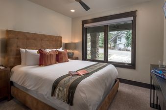 A cozy king bedroom in Frosthaven Chalet features a brown tufted headboard, white bedding with decorative red pillows, and a window offering an outdoor view.