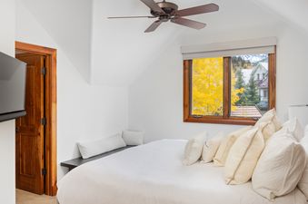 A bright bedroom with a large bed, wooden framed window with a view of autumn trees and mountains, and a ceiling fan.